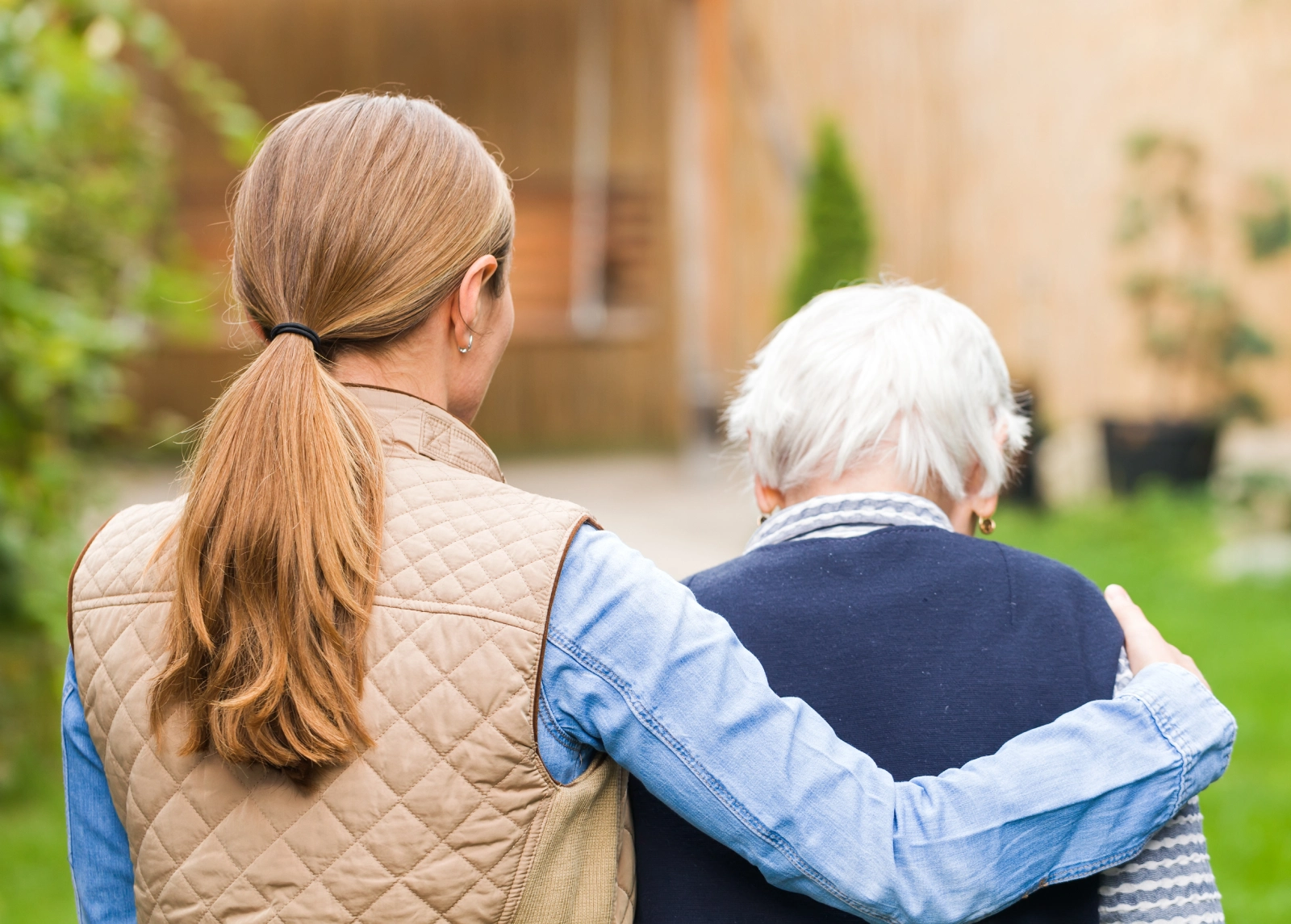 Young woman caring for an elderly woman, walking outdoors with her arm around her shoulder.