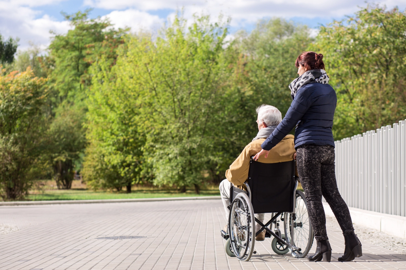 Caregiver pushing an older person in a wheelchair on a paved path outdoors.